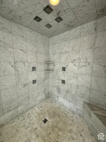 Walk-in shower featuring floor-to-ceiling stone tile, a built-in stone bench, four integrated ceiling shower heads, two wall-mounted shower controls, and two corner storage shelves