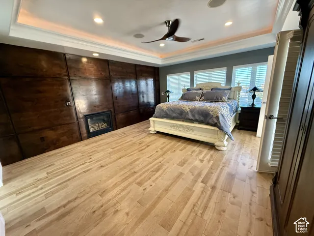 Master bedroom with Tray ceiling with recessed lighting, integrated wood-paneled wall, built-in fireplace, wood-finish flooring, and multi-blade ceiling fan