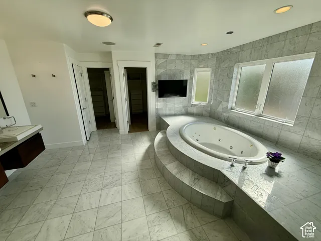 Jetted soaking tub set in a raised stone tile surround, complemented by stone tile flooring and wall cladding, recessed lighting, and a floating vanity