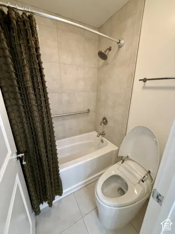 Full bathroom featuring a tub-shower combination with floor-to-ceiling neutral tile surround, wall-mounted safety grab bar, polished chrome fixtures, and a dual-flush toilet