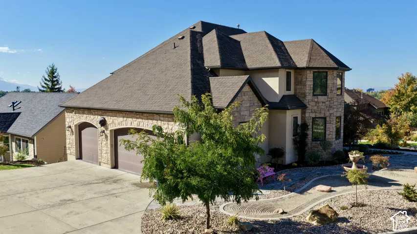 Multi-level exterior featuring a stone facade, dual-bay garage, shingled roof, and landscaped grounds with decorative stone hardscaping