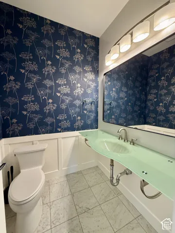 Patterned wallpaper accent wall featuring white wainscoting, glass vanity with an integrated sink, polished metal faucet, and marble tile flooring