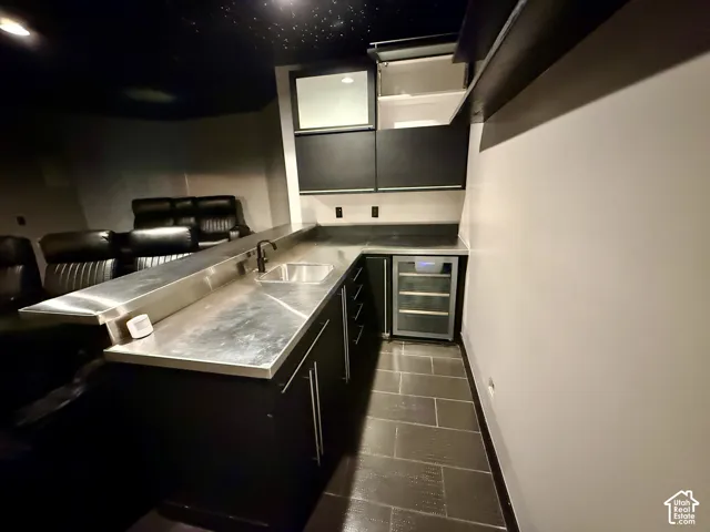 Wet bar featuring custom dark cabinetry, stainless steel countertop, under-mount sink with matte black faucet, integrated beverage cooler, and tile flooring