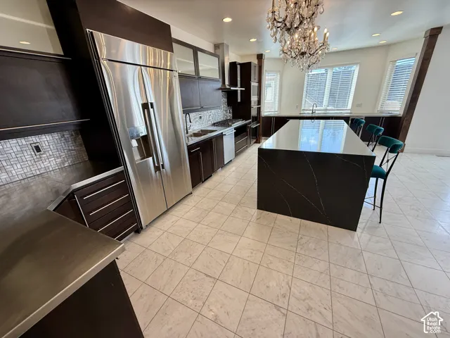 Kitchen featuring dark wood cabinetry, a large central island with a stone countertop, stainless steel refrigerator, tile backsplash.