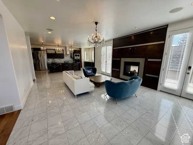 Open-concept space featuring polished stone-finish marble floor, a floor-to-ceiling wood-paneled fireplace surround, integrated recessed lighting, decorative crystal chandeliers, and double French doors