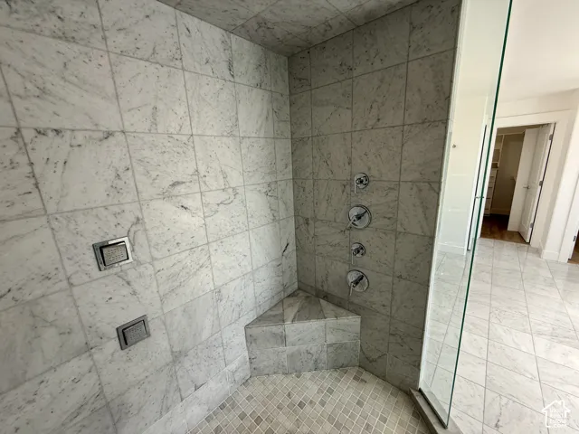 Walk-in shower featuring large-format natural stone wall tiles, a built-in corner bench, multiple chrome wall-mounted fixtures, and a glass partition