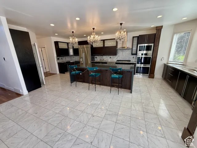 Kitchen featuring dark wood-finish cabinetry, a large central island with an integrated stone countertop, stone tile flooring, stainless steel built-in double wall ovens, and multiple tiered crystal chandeliers