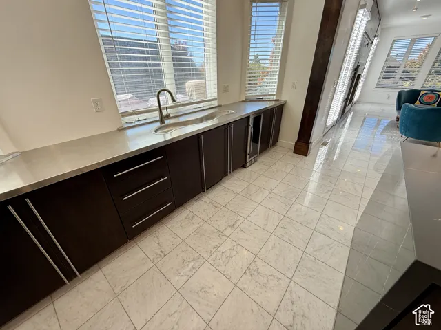 Wet bar featuring dark wood cabinetry, stainless steel countertop with integrated wave sink, built-in under-counter beverage refrigerator, and marble flooring