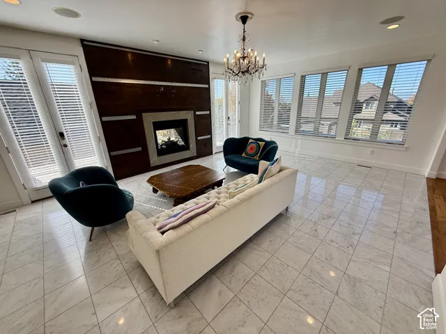 Living area featuring large-format marble flooring, a floor-to-ceiling wood-paneled fireplace surround, a crystal chandelier, multiple floor-to-ceiling windows, and 2 glass-paned doors to the deck and double sided fireplace