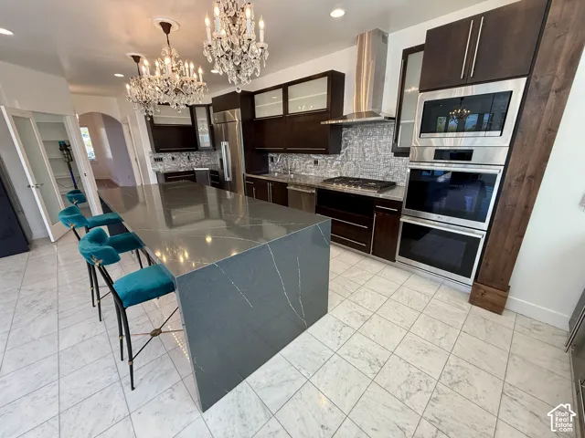 Kitchen featuring a large central island with solid-surface waterfall countertop, dark wood cabinetry, stainless steel double wall ovens, patterned stone tile backsplash, and crystal chandeliers
