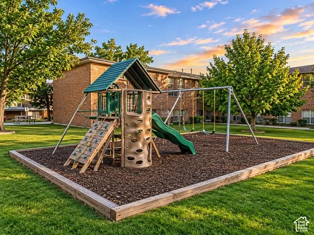 Outdoor recreational area featuring a playground structure with a slide, climbing wall, and swing set, contained within a mulched border on manicured grounds