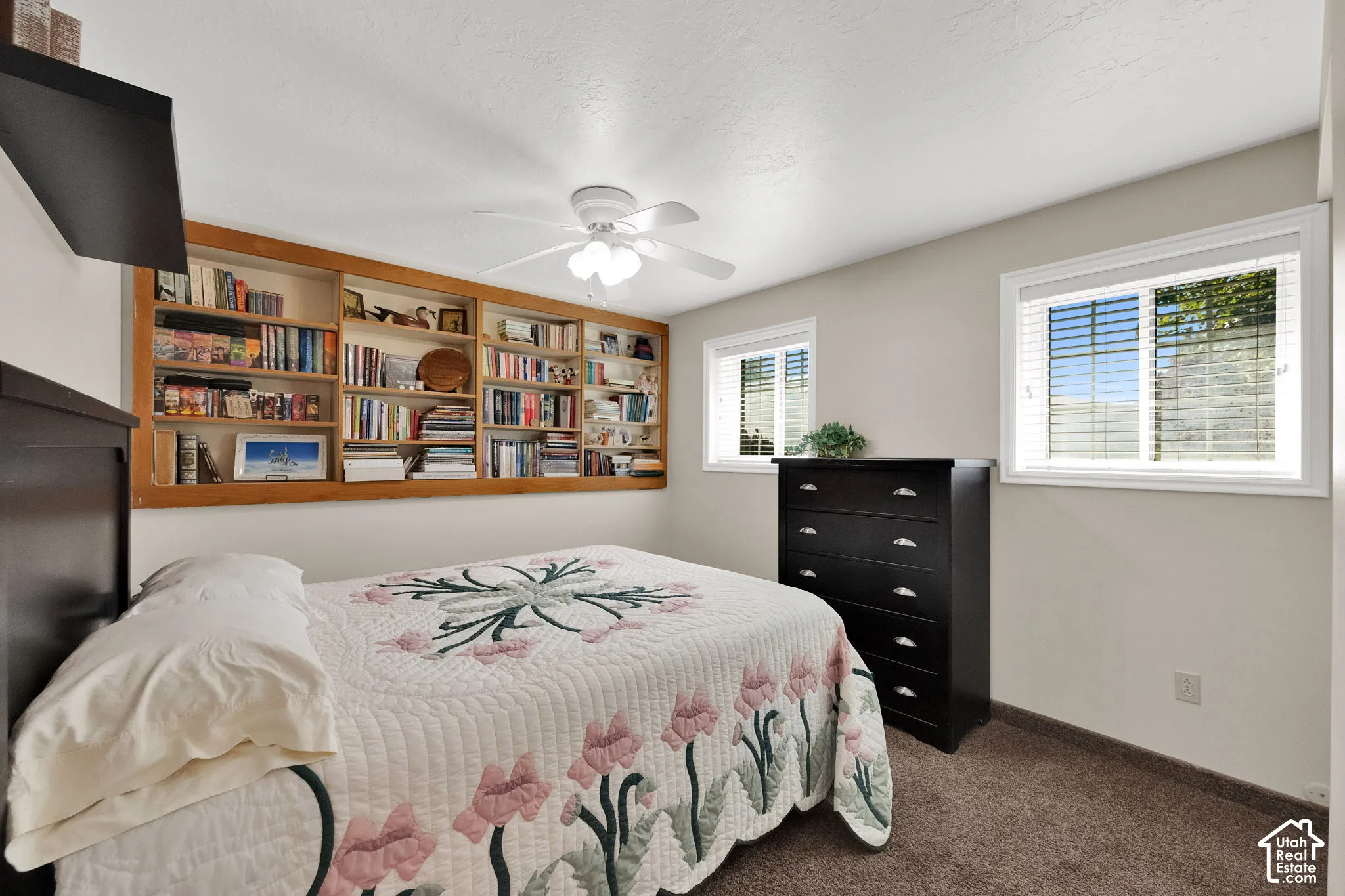 Built-in wood shelving, dual windows with horizontal blinds, neutral carpeted flooring, and a ceiling-mounted light fixture