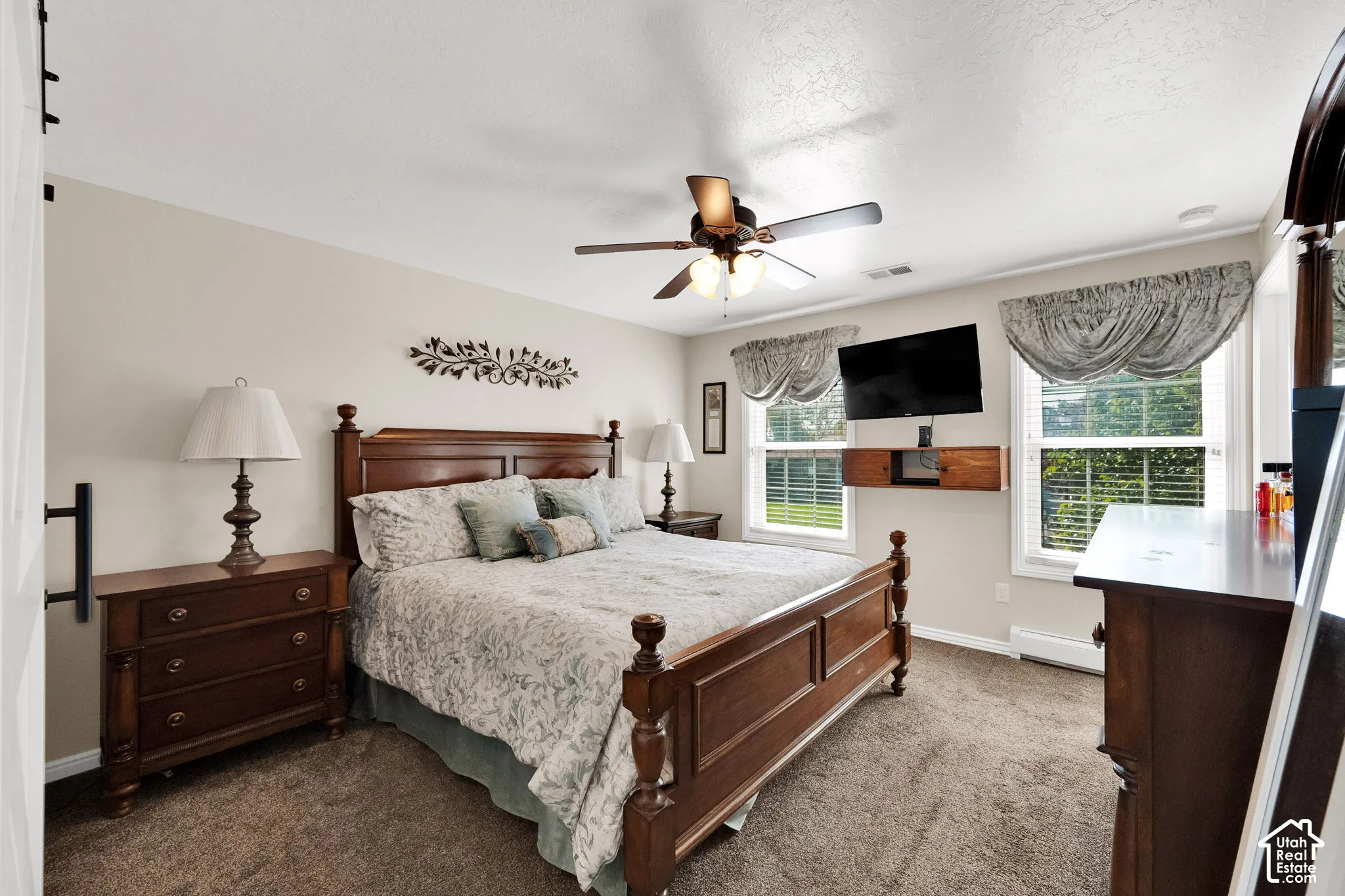 Neutral-toned bedroom featuring wall-to-wall carpeting, a central ceiling fan with integrated lighting, dual windows with decorative treatments, and a wall-mounted floating shelf