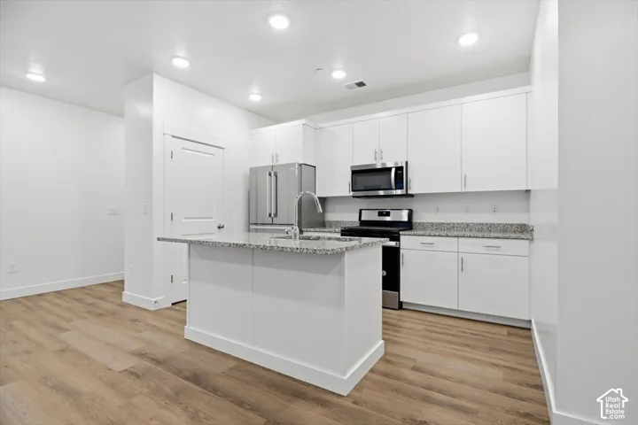 Kitchen featuring white cabinetry, stone-finish countertops, stainless steel appliances, a center island with a sink, and wood-finish flooring