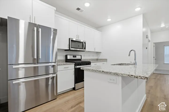 Kitchen featuring white cabinetry, stone-finish countertops, stainless steel appliances, a center island with an integrated sink, and wood-finish flooring