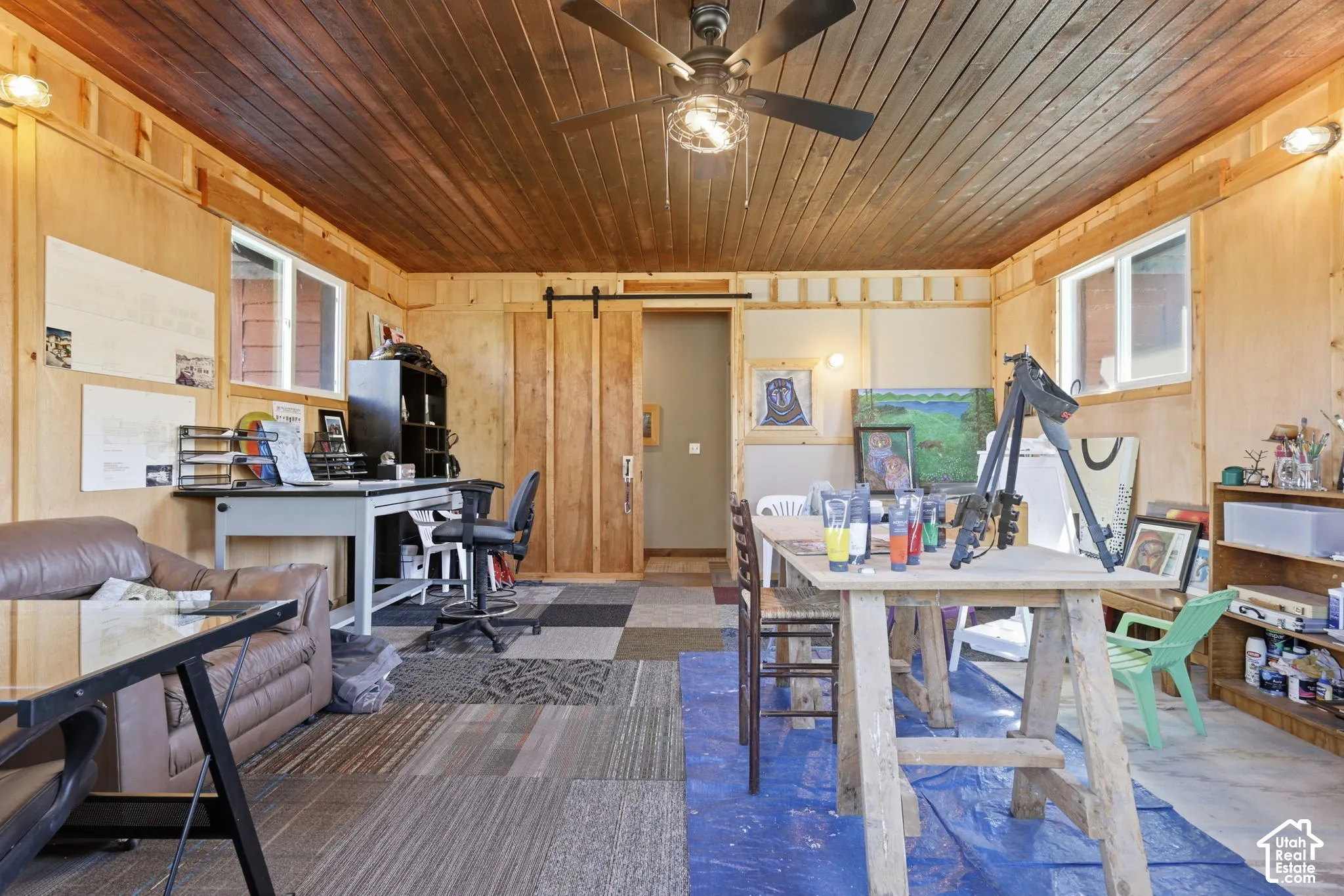 Interior room featuring wood-paneled walls, tongue-and-groove wood ceiling, sliding barn-style door, multi-tone carpet tile flooring, and a ceiling-mounted light fixture with fan