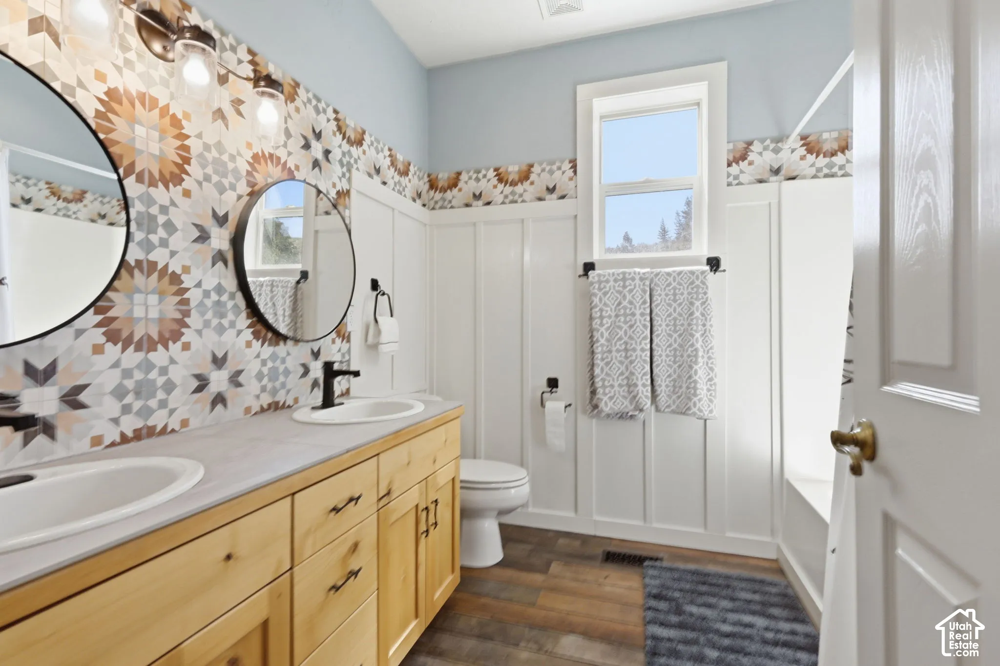 Bathroom featuring a wood-finish vanity with dual undermount sinks, patterned tile backsplash, board and batten wainscoting, and circular wall-mounted mirrors