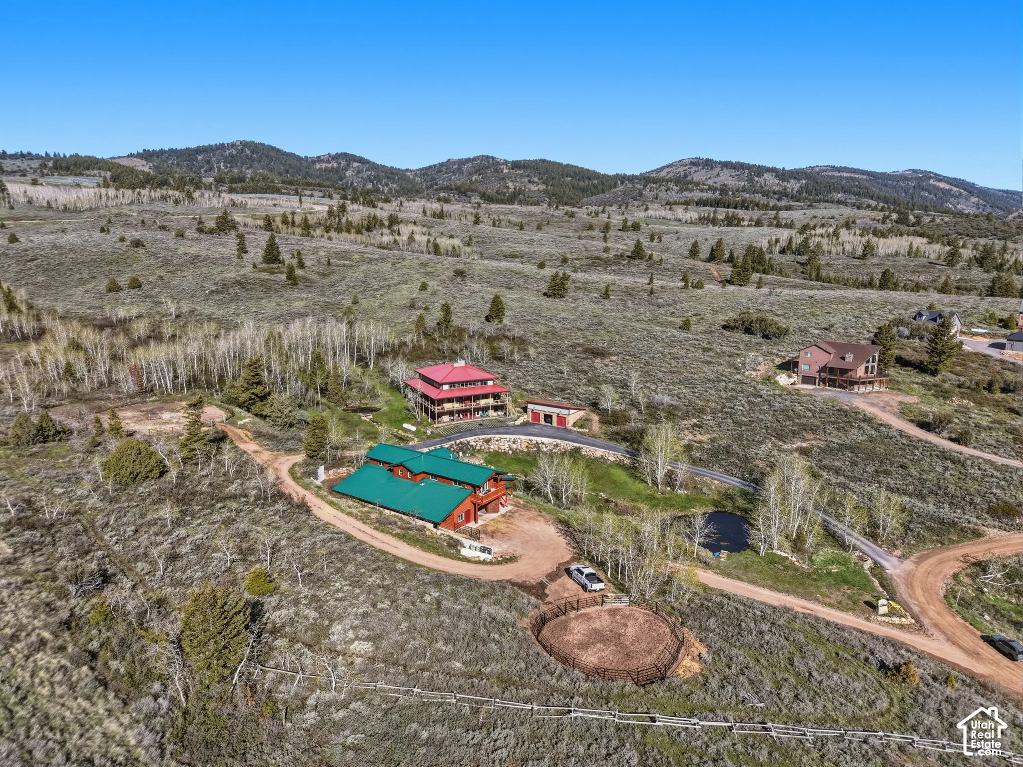 Aerial perspective of a multi-level structure featuring a green metal roof, wrap-around deck, paved driveway, on-site riding arena, and mountain backdrop