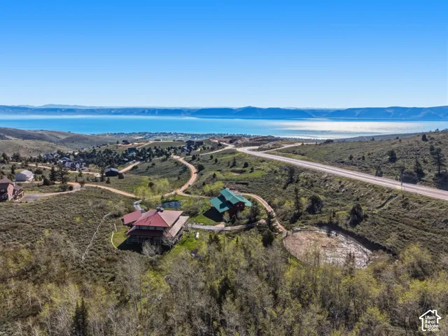 Aerial perspective of a multi-level residence with a metal roof, featuring expansive water and mountain scenery, localized unpaved access paths, and established coniferous and deciduous tree growth