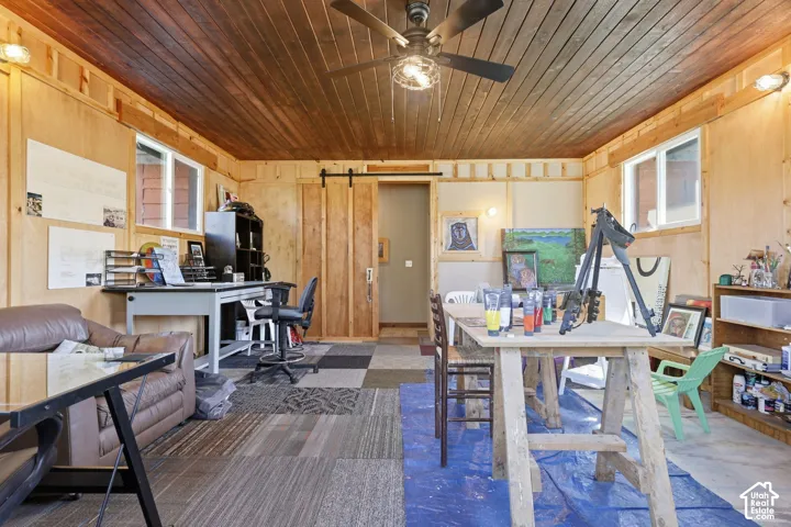 Interior room featuring wood-paneled walls, tongue-and-groove wood ceiling, sliding barn-style door, multi-tone carpet tile flooring, and a ceiling-mounted light fixture with fan