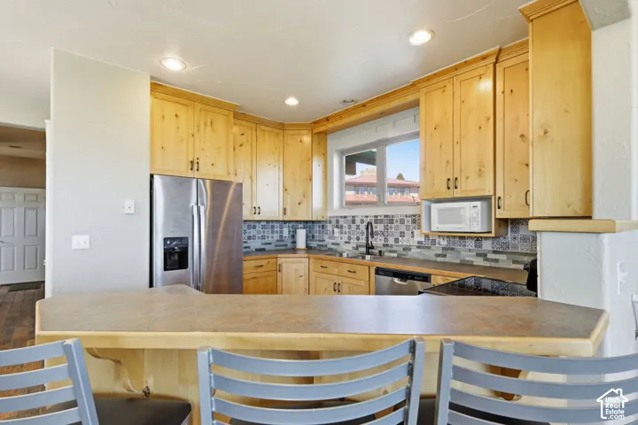 Kitchen featuring natural wood cabinetry, a decorative patterned tile backsplash, stainless steel appliances, a peninsula breakfast bar, and recessed lighting