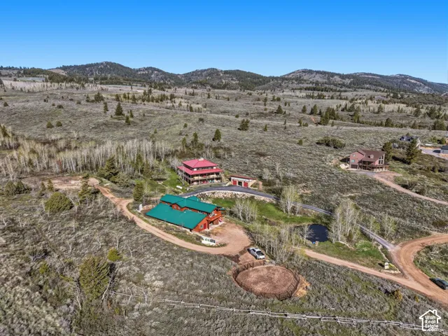Aerial perspective of a multi-level structure featuring a green metal roof, wrap-around deck, paved driveway, on-site riding arena, and mountain backdrop