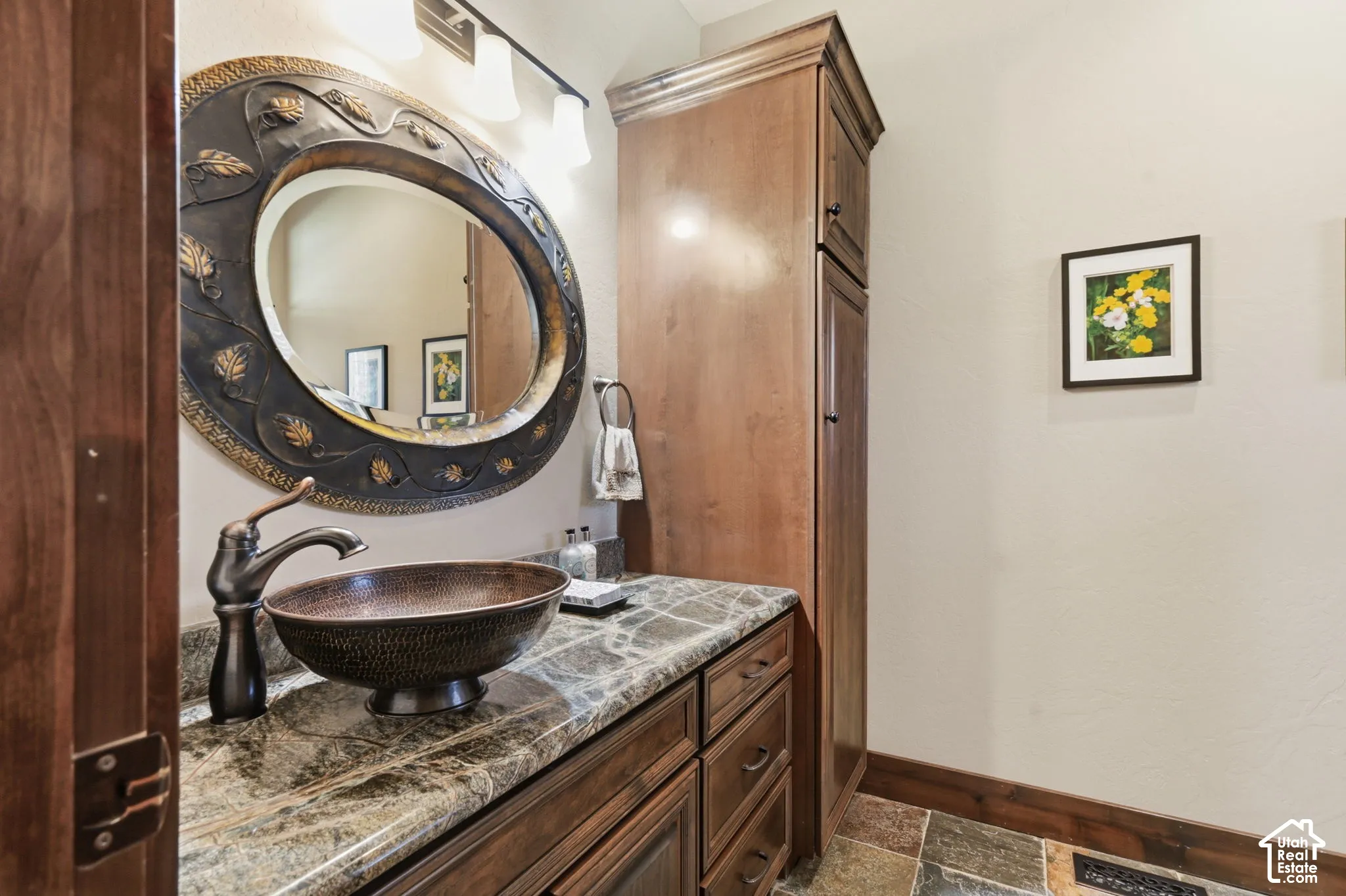Custom wood-finish vanity featuring stone-finish countertop, hammered copper vessel sink, ornate oval mirror, and tall integrated cabinetry