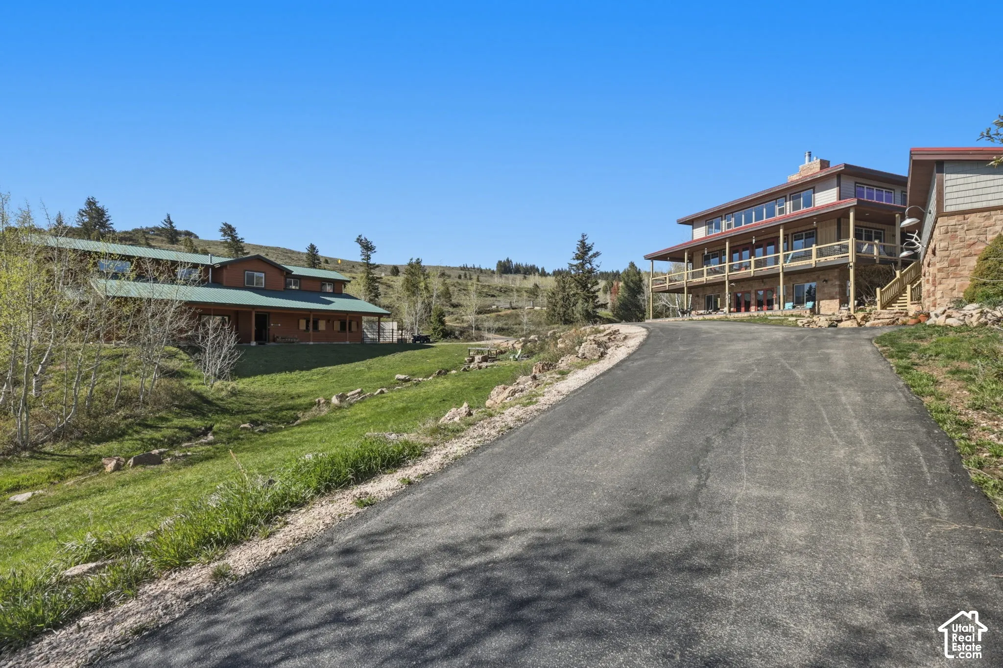 Asphalt driveway leading to a multi-level residence featuring stone and siding exterior, expansive wraparound balconies, and metal roofing, positioned on a large acreage lot with open terrain