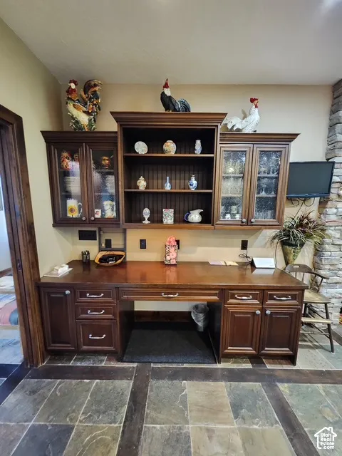 Built-in cabinetry featuring custom woodwork, glass-front upper displays, integrated desk space, and stone tile flooring
