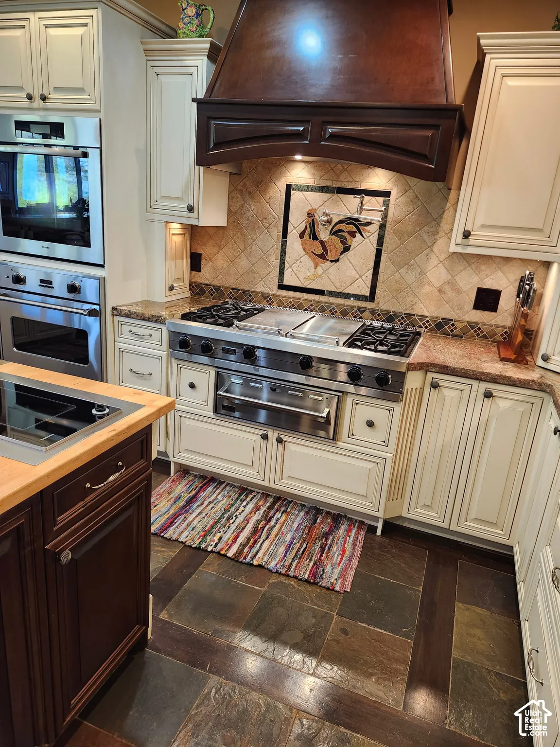 Kitchen featuring custom cabinetry, a multi-burner gas range with integrated stovetop griddle, professional-style stainless steel wall oven, decorative tile backsplash, and stone tile flooring