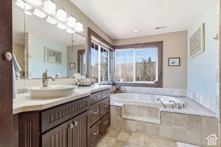 Bathroom featuring a custom dark-wood vanity, vessel sink, stone-tile deck soaking tub, and large picture windows with wood casing