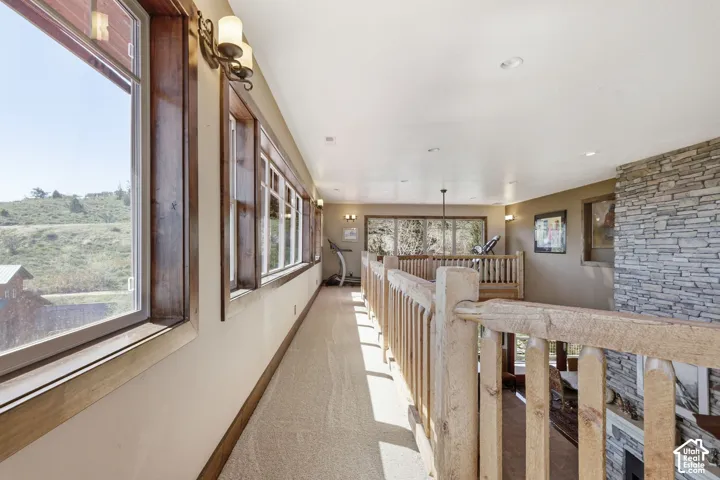 Elevated walkway featuring a stacked stone fireplace surround, natural wood-finish railings, wall-mounted sconce lighting, and expansive windows