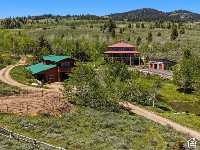 Multi-structure property featuring red exterior siding, metal roofing, a circular metal corral, and a pond, set against a mountainous landscape