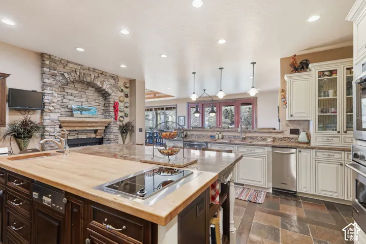 Kitchen featuring a multi-tone island with a wood-finish butcher block surface and integrated cooktop, stone-surround fireplace with arched masonry detail, custom cabinetry, stone-tile flooring, and recessed lighting