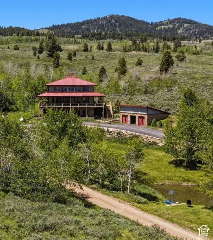 Multi-level residence featuring a red metal roof, expansive wrap-around wooden decking, stone foundation, and a detached two-car garage, set against a mountainous backdrop with a private pond