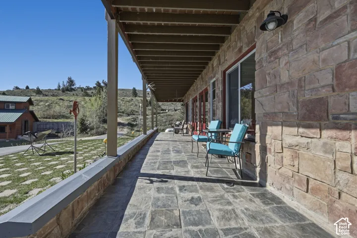 Covered stone-paved patio featuring a natural stone veneer exterior, exposed wood-beam ceiling, wall-mounted exterior lighting, and sliding glass doors