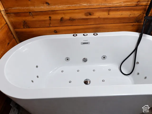 Freestanding jetted soaking tub featuring chrome hydrotherapy jets, a center-mount chrome faucet, a flexible black hand-held shower wand, and natural wood-plank wall paneling