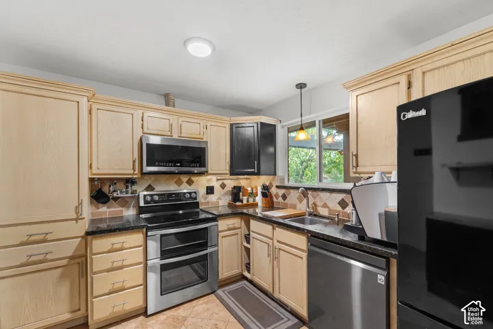 Kitchen featuring light-tone wood cabinetry, dark stone-finish countertops, stainless steel appliances, and multi-toned tile backsplash