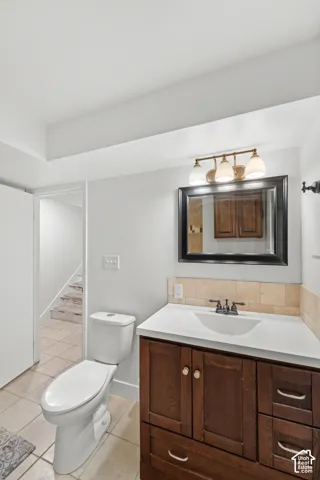 Bathroom featuring a wood-finish vanity with an integrated basin, natural stone tile backsplash, wall-mounted framed mirror, three-light vanity fixture, and neutral ceramic tile flooring