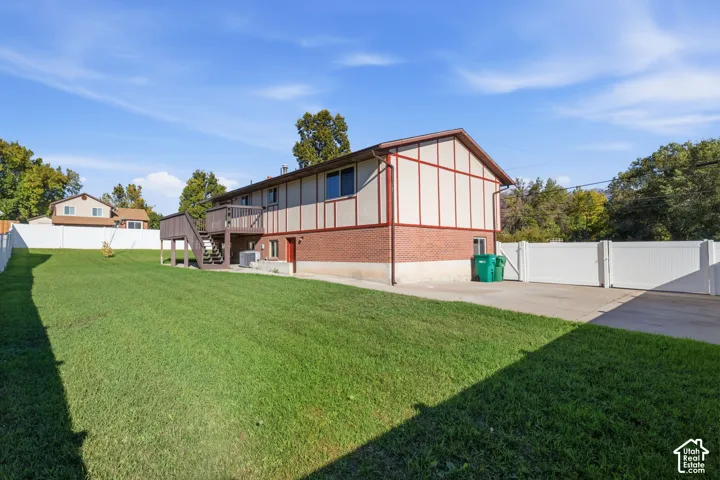 Expansive backyard featuring a manicured lawn, elevated wood deck with stairs, concrete driveway, and white vinyl privacy fencing