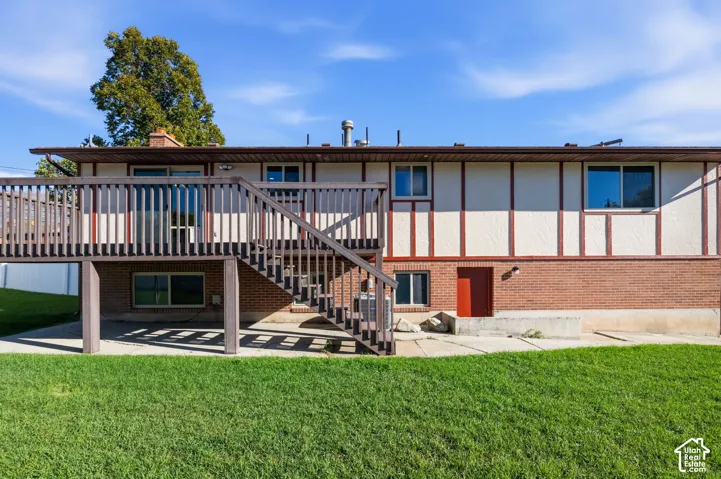 Raised wood deck with staircase, brick and stucco exterior, paved ground-level patio, and lawn