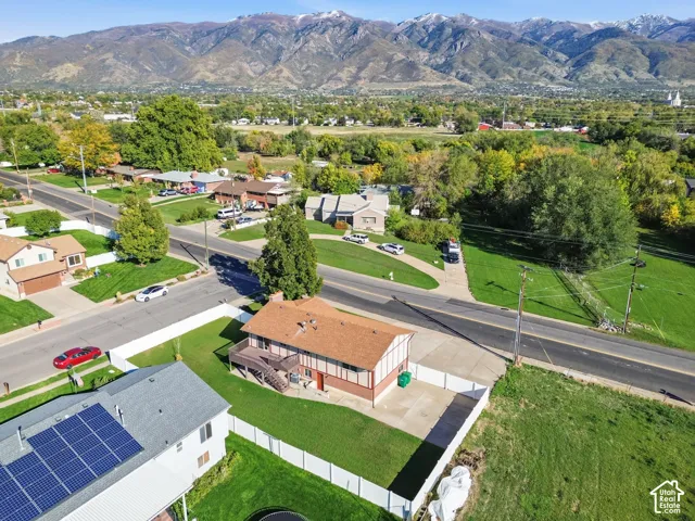 Aerial perspective featuring a multi-level residence with an elevated deck, attached staircase, concrete parking area, and a white privacy fence, set against a prominent mountain backdrop
