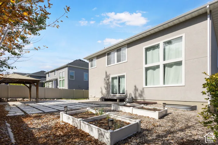 Rear exterior featuring a neutral stucco facade, large windows, sliding glass door, paved stone patio, and raised garden beds with a perimeter vinyl fence
