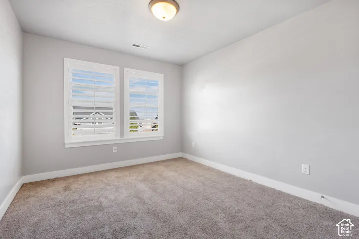 Neutral-toned interior space featuring light grey walls, carpeted flooring, two windows with white plantation shutters, and a central ceiling-mounted light fixture