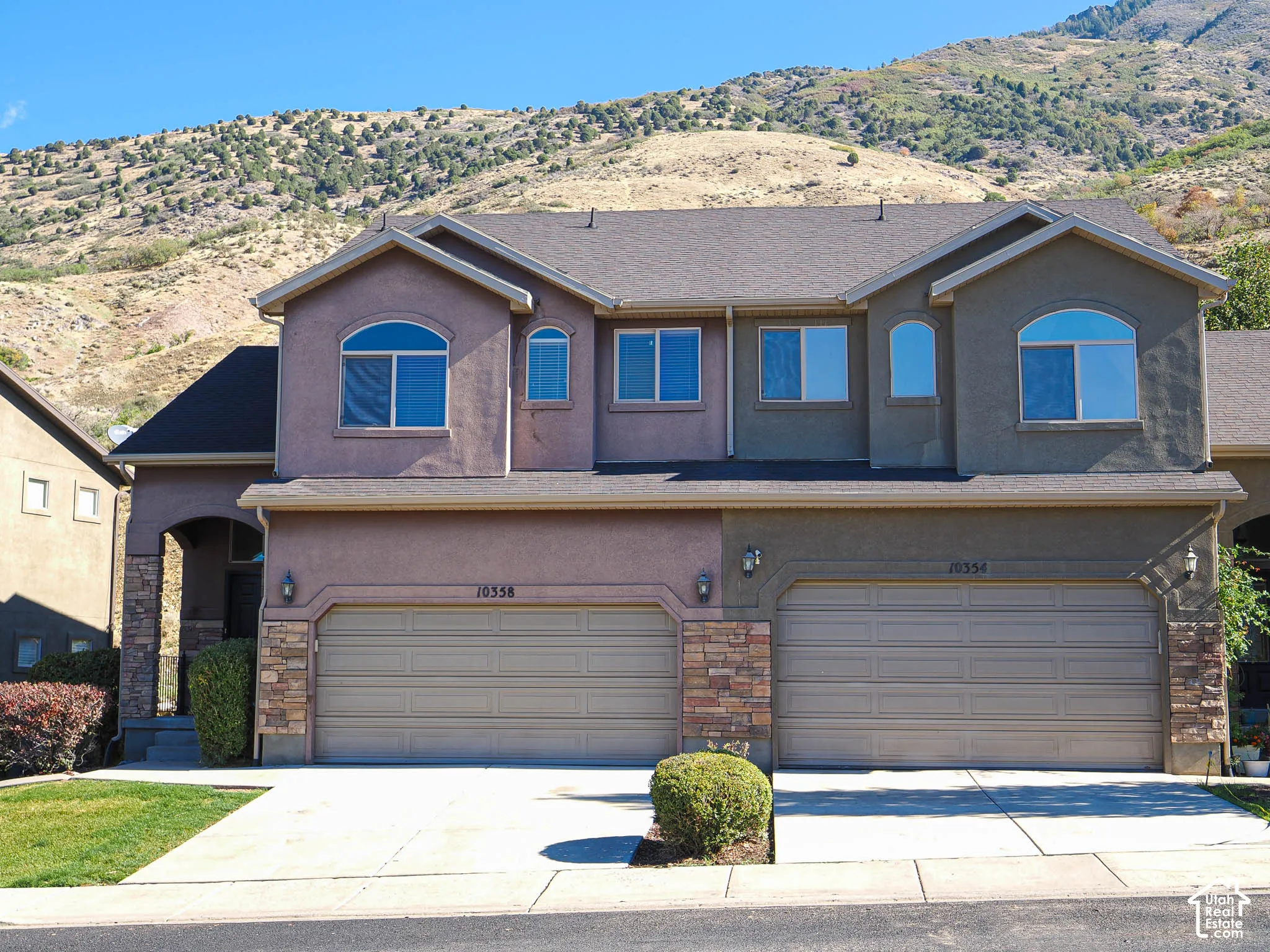 Two-story stucco exterior featuring stone veneer accents, multi-level rooflines, dual double-car garages, and arched window details, set against a mountainous backdrop