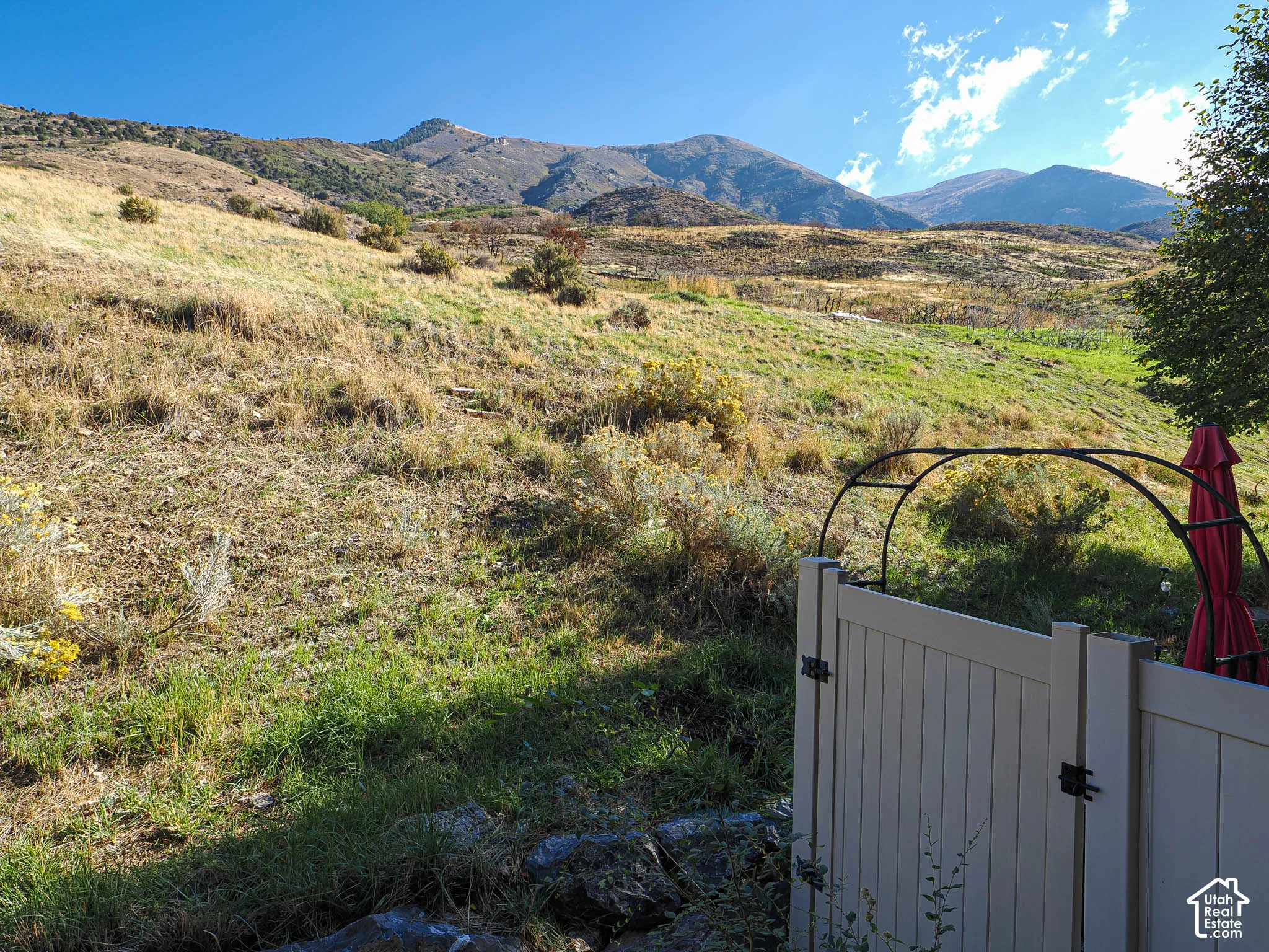 Expansive hillside landscape featuring natural terrain and a mountain backdrop, complemented by a white vinyl privacy fence and a metal garden arch