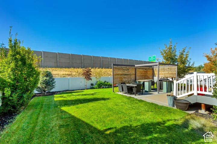 Backyard featuring a manicured lawn, concrete patio, custom metal and wood-slat pergola, white vinyl perimeter fencing, and mature landscaping
