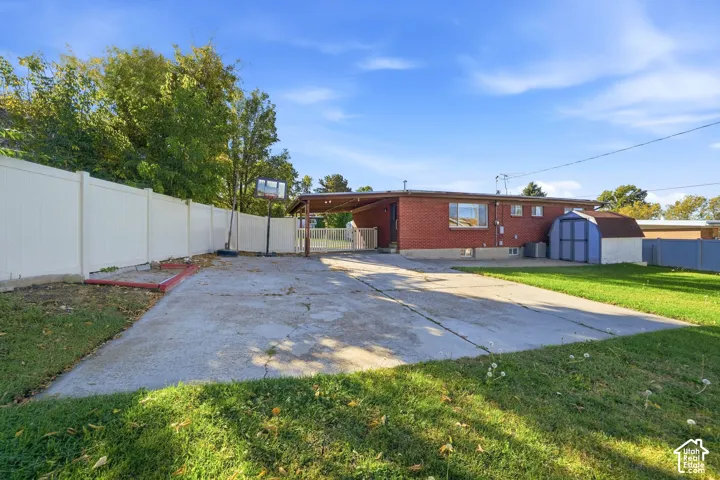 Red brick exterior featuring a covered carport, expansive concrete driveway, vinyl privacy fencing, and a detached storage shed