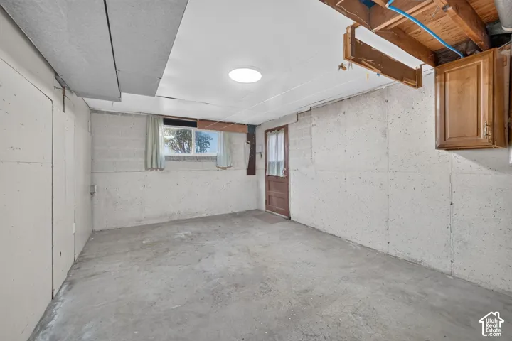 Unfinished storage area featuring exposed wood ceiling joists, concrete block walls, concrete flooring, built-in wooden cabinetry, and an exterior door