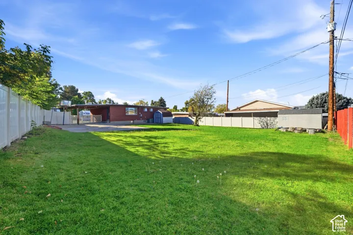 Expansive backyard featuring a large grass lawn, perimeter vinyl fencing, a paved parking area, and a detached storage structure